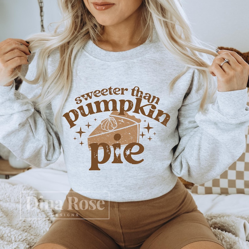 Pode incluir: Uma camisa de moletom cinza com o texto "sweeter than pumpkin pie" em letras marrons. Uma imagem estilizada de uma fatia de torta de ab&oacute;bora est&aacute; no centro do texto.