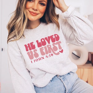 May include: A gray sweatshirt with the text "He Loved Us First 1 John 4:19" in pink and red lettering. The text is in a retro font style with a heart symbol.