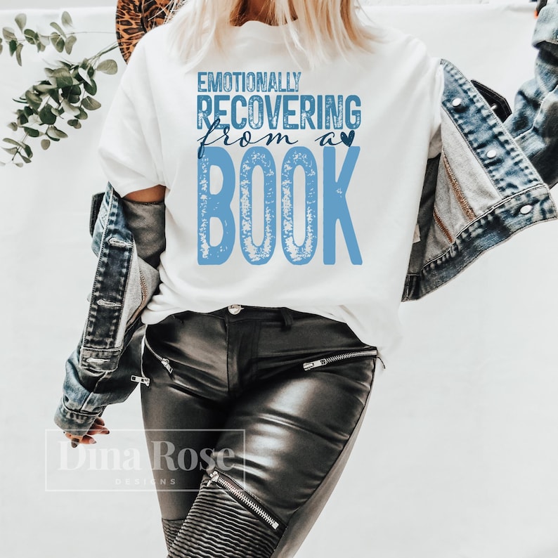 May include: White t-shirt with the text "Emotionally Recovering From a Book" in blue. The shirt is worn with a denim jacket and black leather trousers. The design features a heart symbol above the word "book".