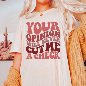 May include: A white t-shirt with the text "YOUR OPINION WILL NEVER CUT ME A CHECK" in a retro, colorful font.