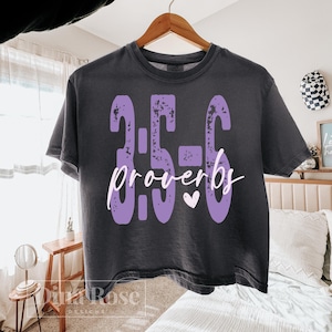 May include: Dark gray t-shirt with the text "3:5-6 Proverbs" in purple, featuring a distressed font and a small white heart. The shirt is hanging on a wooden hanger.