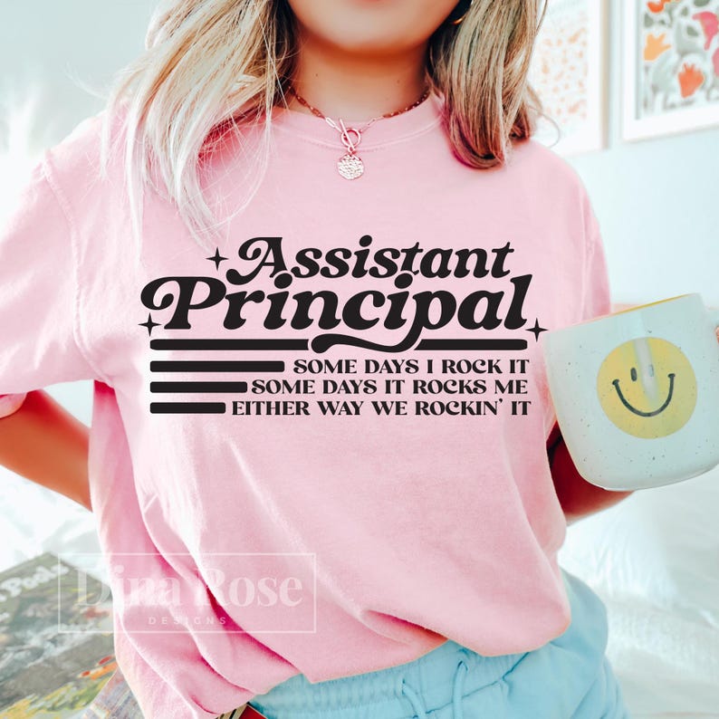 May include: A light pink t-shirt with the words "Assistant Principal" in a black, stylized font. Below the title, the shirt reads "SOME DAYS I ROCK IT, SOME DAYS IT ROCKS ME, EITHER WAY WE ROCKIN' IT." A white mug with a yellow smiley face is visible.