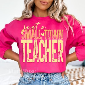 Pu&ograve; includere: Una felpa rosa acceso con la scritta "just a SMALL-TOWN TEACHER" in giallo. Il testo ha un simbolo a cuore tra "SMALL" e "TOWN". La felpa &egrave; a girocollo ed &egrave; realizzata in un materiale morbido.