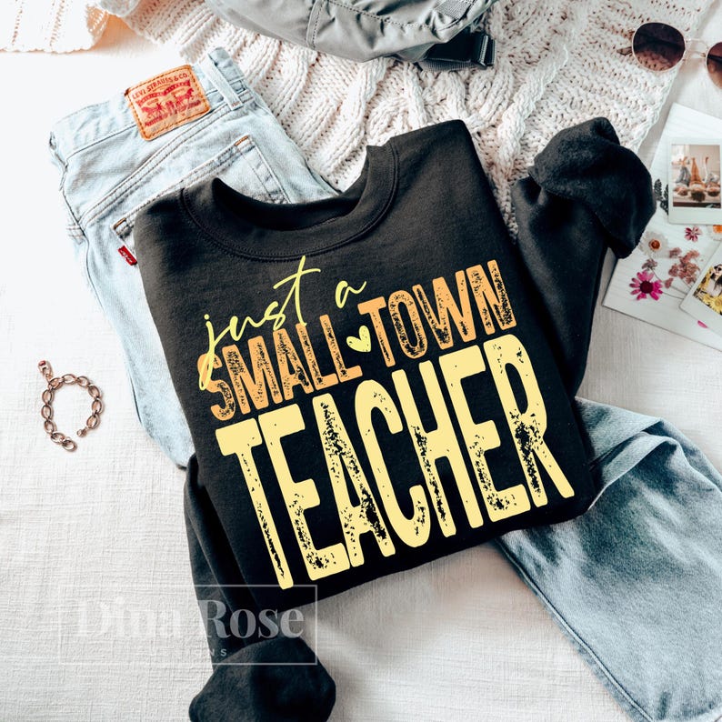 May include: Black crewneck sweatshirt with the text "just a SMALL TOWN TEACHER" in yellow and orange. The sweatshirt is laid on top of light wash denim jeans. A silver bracelet and sunglasses are also visible.