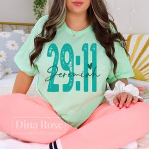 May include: Mint green t-shirt with the text "29:11 Jeremiah" in teal. The shirt is paired with pink pants. The Dina Rose Designs logo is visible at the bottom left.