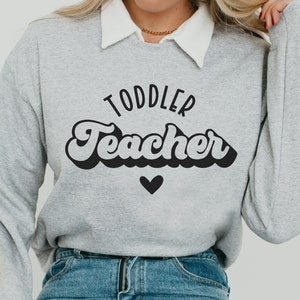 Pu&ograve; includere: Una felpa grigia con il testo "Toddler Teacher" in un font retr&ograve; con un cuore nero sotto il testo.