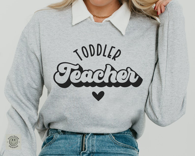 Pu&ograve; includere: Una felpa grigia con il testo "Toddler Teacher" in un font retr&ograve; con un cuore nero sotto il testo.