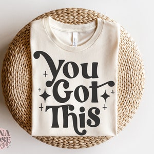 Puede incluir: Una camiseta beige con el texto "You Got This" en negro con una fuente retro y peque&ntilde;as estrellas brillantes.