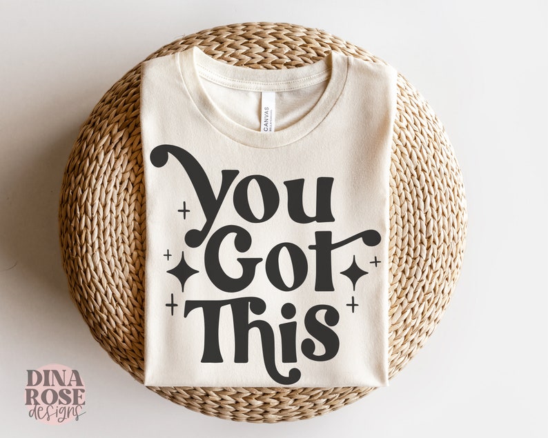 Puede incluir: Una camiseta beige con el texto "You Got This" en negro con una fuente retro y peque&ntilde;as estrellas brillantes.