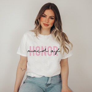 May include: White t-shirt with the text "maid of honor" in pink lettering.