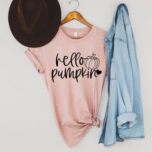 May include: A pink t-shirt with the text "hello pumpkin" and a small pumpkin graphic. The shirt is hanging on a wooden hanger with a denim jacket draped over it.