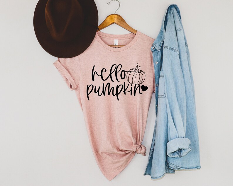 May include: A pink t-shirt with the text "hello pumpkin" and a small pumpkin graphic. The shirt is hanging on a wooden hanger with a denim jacket draped over it.