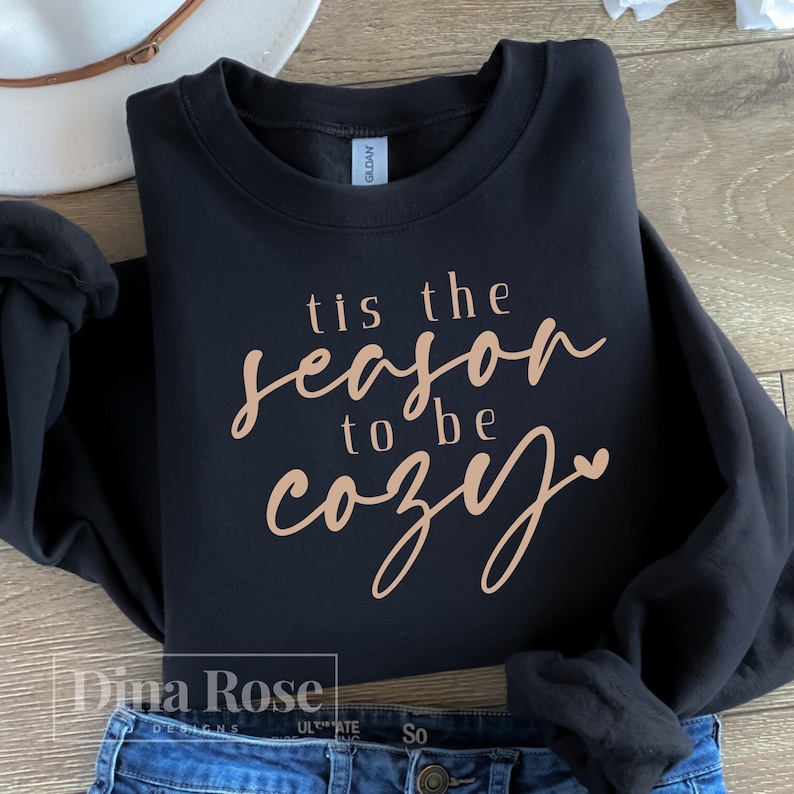 Puede incluir: Sudadera negra con la frase "tis the season to be cozy" en fuente cursiva. El texto es de color marr&oacute;n claro. La sudadera est&aacute; doblada sobre unos vaqueros azules. Un sombrero blanco es visible en el fondo.