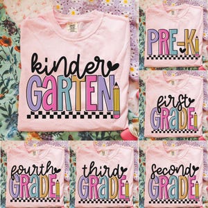 Puede incluir: Camisetas rosas con diseños de niveles escolares. Las camisetas presentan las palabras "Kindergarten", "Pre-K", "First Grade", "Fourth Grade", "Third Grade" y "Second Grade" en fuentes coloridas con gráficos de lápices.