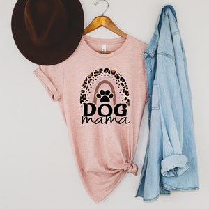 May include: A pink t-shirt with a leopard print rainbow and paw print design. The text "DOG mama" is printed below the rainbow. The shirt is hanging on a wooden hanger with a denim jacket and a brown hat.