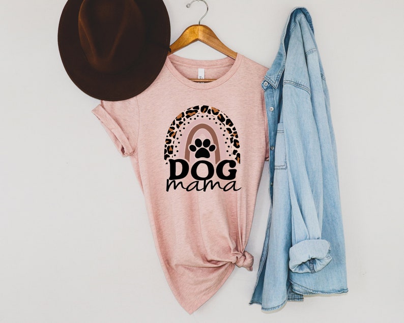 May include: A pink t-shirt with a leopard print rainbow and paw print design. The text "DOG mama" is printed below the rainbow. The shirt is hanging on a wooden hanger with a denim jacket and a brown hat.