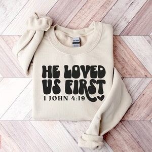 May include: A beige sweatshirt with the text "He Loved Us First" in black, with a heart and the scripture reference "1 John 4:19" underneath.