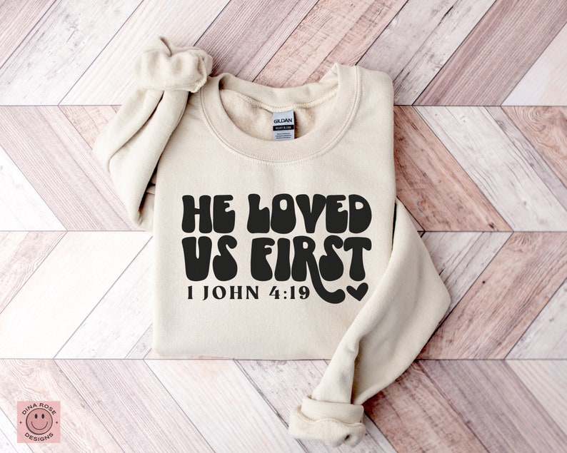 May include: A beige sweatshirt with the text "He Loved Us First" in black, with a heart and the scripture reference "1 John 4:19" underneath.