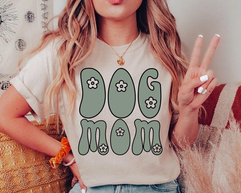 May include: A white t-shirt with a green retro-style graphic that says "Dog Mom" with small white flowers in the letters.