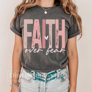 May include: Dark gray t-shirt with the words "FAITH over fear" in pink and white lettering. The word "FAITH" is in a bold, blocky font, and the word "fear" is in a cursive font. A small heart is between the words.