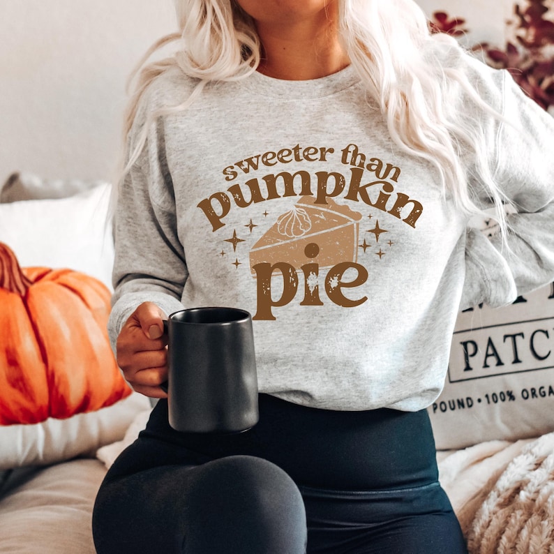 Pode incluir: Uma camisa de moletom cinza com o texto "sweeter than pumpkin pie" em letras marrons. O texto &eacute; cercado por estrelas e uma ilustra&ccedil;&atilde;o marrom de uma fatia de torta de ab&oacute;bora.