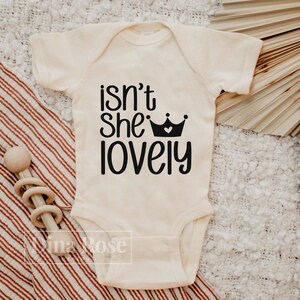 May include: A white baby onesie with black text that reads "isn't she lovely" with a black crown with a white heart in the center.
