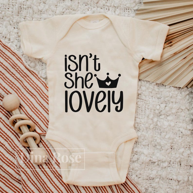 May include: A white baby onesie with black text that reads "isn't she lovely" with a black crown with a white heart in the center.