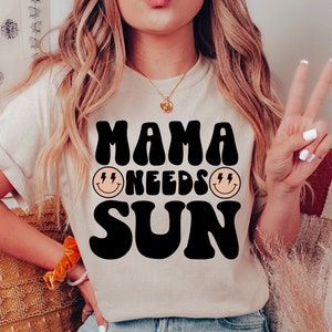 May include: A white t-shirt with black text that reads "Mama Needs Sun" in a retro font. The text is surrounded by two yellow smiley faces with lightning bolts for eyes.