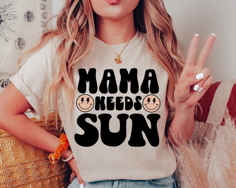 May include: A white t-shirt with black text that reads "Mama Needs Sun" in a retro font. The text is surrounded by two yellow smiley faces with lightning bolts for eyes.
