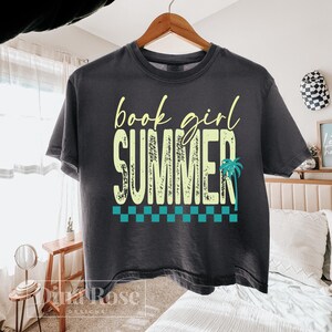 May include: Dark gray t-shirt with the words "book girl SUMMER" in yellow and a teal palm tree graphic. The shirt has a checkered pattern at the bottom and is hanging on a wooden hanger.