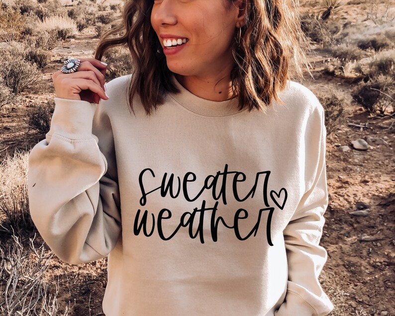 Puede incluir: Una sudadera beige con el texto "sweater weather" en letra negra con un peque&ntilde;o coraz&oacute;n.