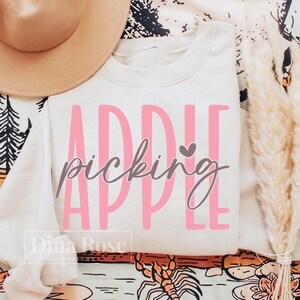 May include: A cream-colored crewneck sweatshirt featuring the pink text APPLE and the brown cursive script picking with a small heart icon. The design is centered on the front of the garment, which is displayed alongside a tan hat and dried grass.