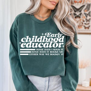 以下が含まれることがあります： 「Early childhood educator」の白い文字と、「Some days I rock it」、「Some days it rocks me」、「Either way we rockin' it」というフレーズがプリントされた、濃いティール色のスウェットシャツ。
