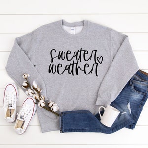 Puede incluir: Una sudadera gris con el texto "sweater weather" en escritura negra con un peque&ntilde;o coraz&oacute;n. La sudadera est&aacute; colocada sobre una superficie de madera blanca con un par de zapatillas blancas, una rama de algod&oacute;n y un par de pantalones vaqueros azules.