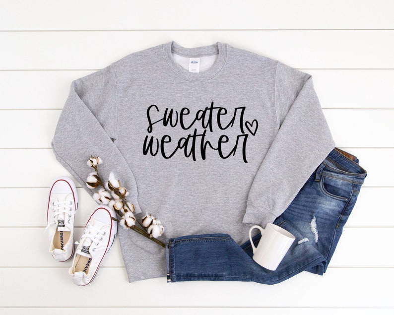 Puede incluir: Una sudadera gris con el texto "sweater weather" en escritura negra con un peque&ntilde;o coraz&oacute;n. La sudadera est&aacute; colocada sobre una superficie de madera blanca con un par de zapatillas blancas, una rama de algod&oacute;n y un par de pantalones vaqueros azules.