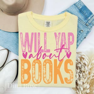 May include: A pale yellow t-shirt with the slogan "WILL YAP about BOOKS" in pink and orange. The shirt is folded on top of blue denim jeans, with a hat and white boots in the background.