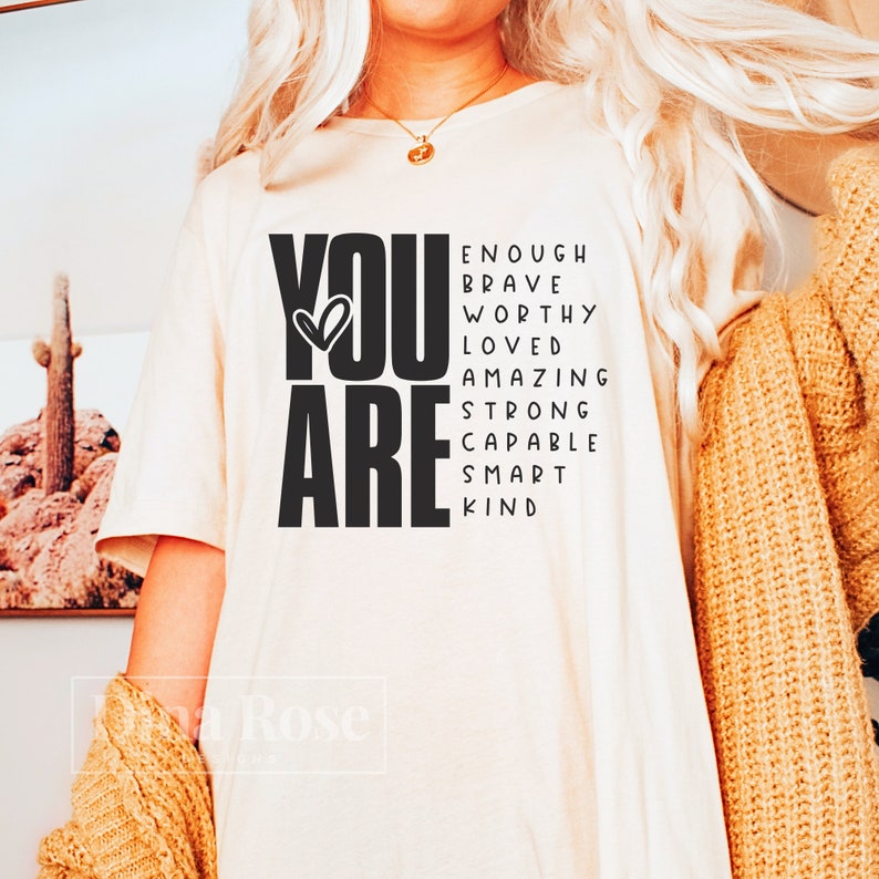 May include: A white t-shirt with the words "YOU ARE" in black, with a heart symbol in the "O". The words "ENOUGH, BRAVE, WORTHY, LOVED, AMAZING, STRONG, CAPABLE, SMART, KIND" are arranged around the words "YOU ARE" in a word search style.