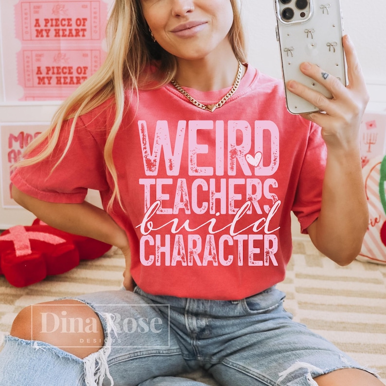 Pu&ograve; includere: Una maglietta rossa con la scritta "WEIRD TEACHERS build CHARACTER" in rosa. La maglietta &egrave; indossata da una persona che indossa jeans blu. L'immagine mostra anche una collana d'oro e un telefono.