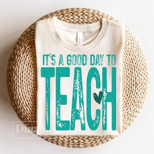 Pu&ograve; includere: T-shirt color crema con la scritta turchese "It's a good day to teach". La parola "teach" &egrave; grande, con un piccolo cuore. La maglietta &egrave; piegata e posizionata su una superficie circolare intrecciata.