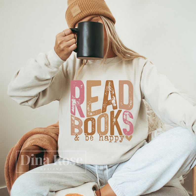 May include: A beige sweatshirt with the text "READ BOOKS & be happy" in pink, brown, and orange letters with a distressed look. The text is centered on the sweatshirt.