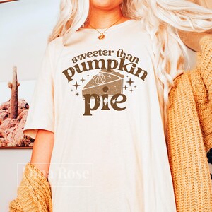 Pode incluir: Uma camiseta branca com o texto "sweeter than pumpkin pie" em letras marrons. O texto &eacute; cercado por estrelas e uma ilustra&ccedil;&atilde;o marrom de uma torta de ab&oacute;bora.