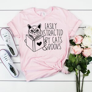 K&ouml;nnte beinhalten: Rosa T-Shirt mit einer schwarzen Illustration einer Katze, die ein Buch liest, mit dem Text "Easily Distracted by Cats & Books".