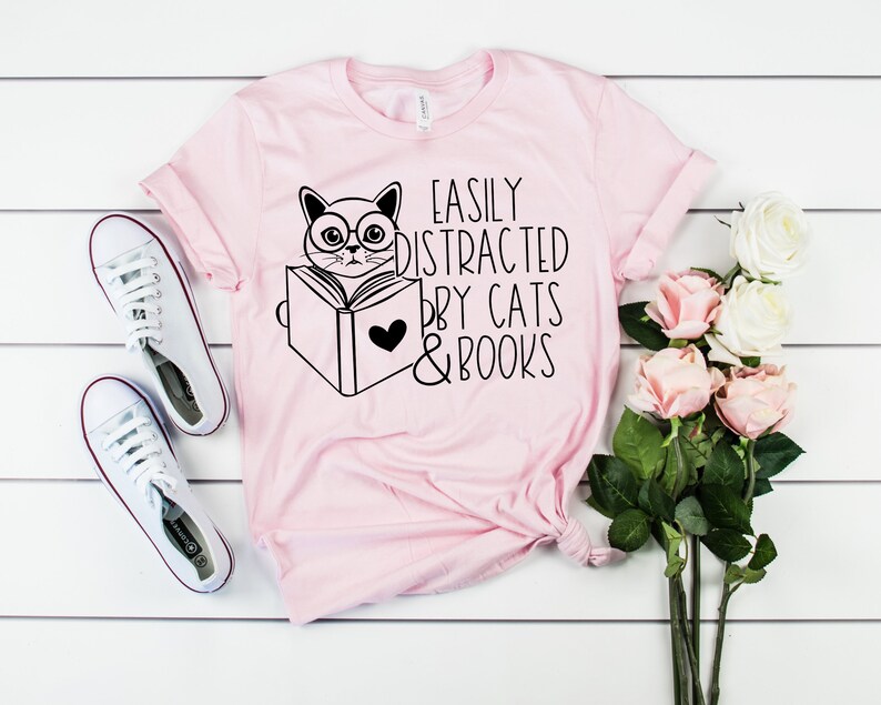 K&ouml;nnte beinhalten: Rosa T-Shirt mit einer schwarzen Illustration einer Katze, die ein Buch liest, mit dem Text "Easily Distracted by Cats & Books".