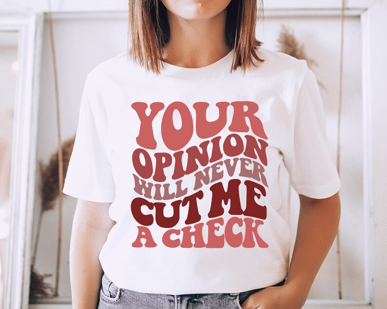 May include: White t-shirt with a pink and red graphic that says "Your opinion will never cut me a check."