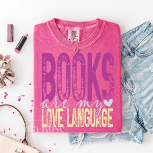 May include: A pink t-shirt with the text "BOOKS are my LOVE LANGUAGE" in purple and yellow. The shirt is folded and placed on a white surface next to denim shorts, a necklace, lipstick, and a scrunchie. A small heart graphic is included.