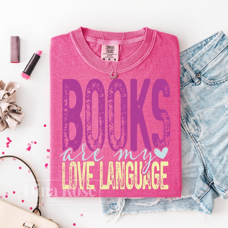 May include: A pink t-shirt with the text "BOOKS are my LOVE LANGUAGE" in purple and yellow. The shirt is folded and placed on a white surface next to denim shorts, a necklace, lipstick, and a scrunchie. A small heart graphic is included.