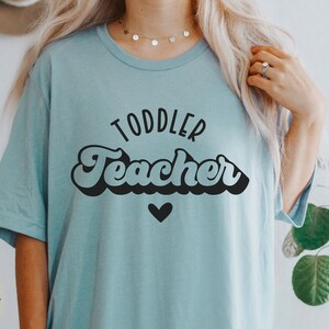 Pu&ograve; includere: Una maglietta blu chiaro con il testo "Toddler Teacher" in un font retr&ograve;. Il testo &egrave; in nero e c'&egrave; un piccolo cuore nero sotto.