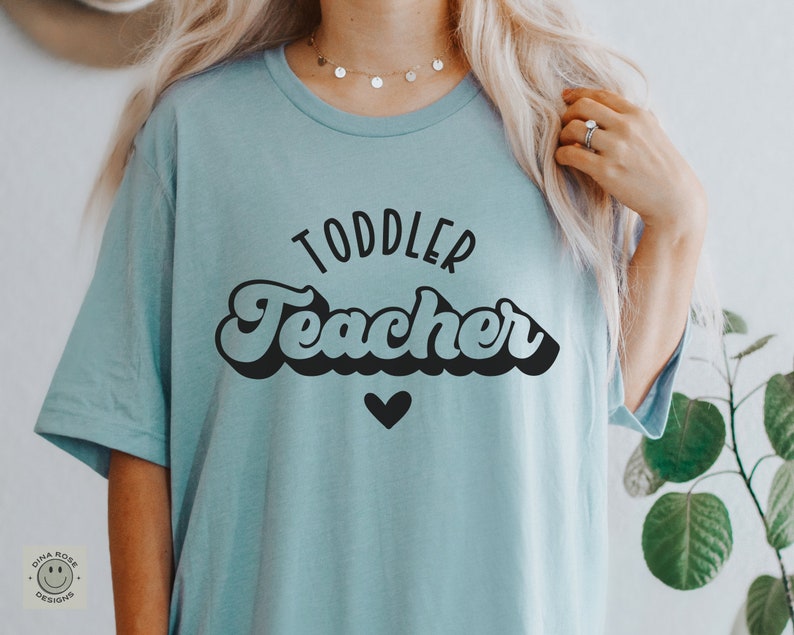 Pu&ograve; includere: Una maglietta blu chiaro con il testo "Toddler Teacher" in un font retr&ograve;. Il testo &egrave; in nero e c'&egrave; un piccolo cuore nero sotto.