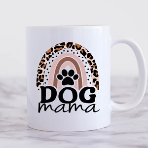 May include: White ceramic coffee mug with a rainbow design featuring a paw print in the center. The rainbow is made of leopard print and the text "Dog mama" is printed below the rainbow.