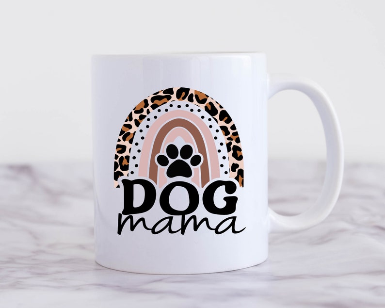 May include: White ceramic coffee mug with a rainbow design featuring a paw print in the center. The rainbow is made of leopard print and the text "Dog mama" is printed below the rainbow.